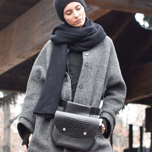 May include: A woman wearing a grey wool coat with a black knitted scarf and a black leather belt bag. The bag has a crocodile-embossed design and a flap closure with silver poppers.