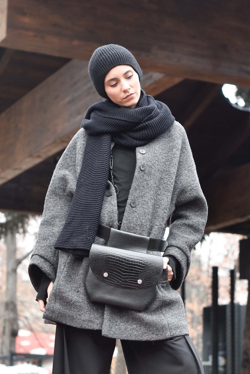 May include: A woman wearing a grey wool coat with a black knitted scarf and a black leather belt bag. The bag has a crocodile-embossed design and a flap closure with silver poppers.