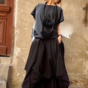 May include: A woman wearing a black, loose-fitting top and skirt with a black rope necklace with black pom-poms. She is wearing black sunglasses and black heeled sandals.