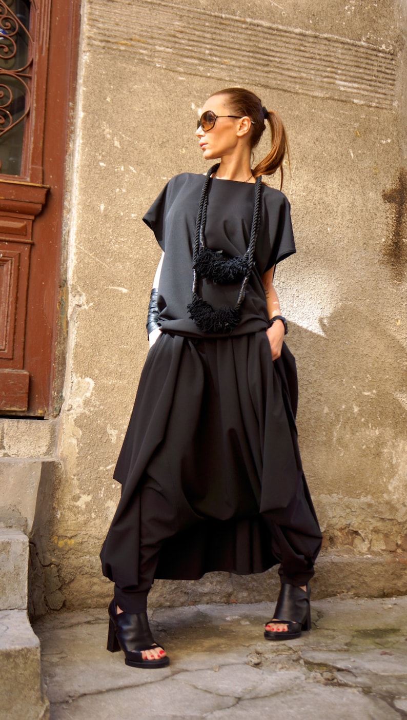 May include: A woman wearing a black, loose-fitting top and skirt with a black rope necklace with black pom-poms. She is wearing black sunglasses and black heeled sandals.