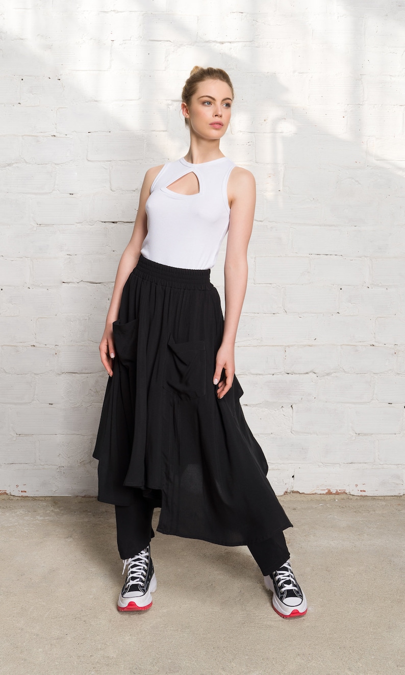 Pode incluir: Uma mulher vestindo um top branco sem mangas com um decote de fechadura e uma saia preta assim&eacute;trica com bolsos. Ela est&aacute; usando t&ecirc;nis preto e branco com detalhes vermelhos.