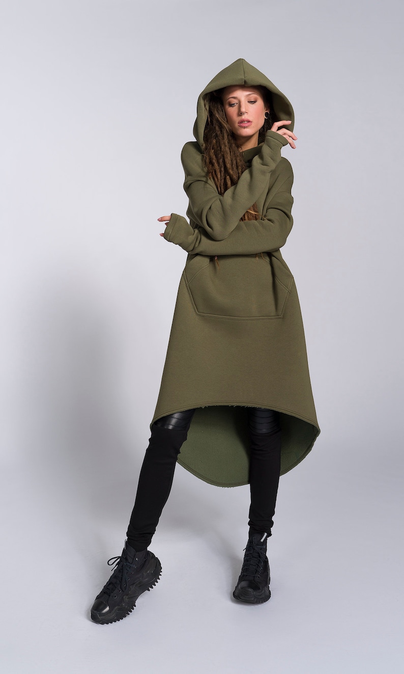 May include: A long, olive green hooded sweatshirt with a high-low hemline. The sweatshirt has a large kangaroo pocket and long sleeves. The model is wearing black leggings and black high-top sneakers.