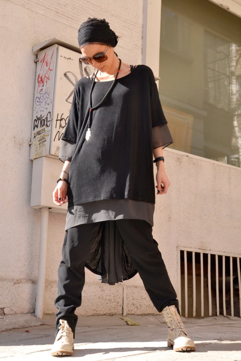 May include: A woman wearing a black long-sleeved shirt with a sheer overlay, black pants, and a black headscarf. She is wearing a silver necklace with a charm and a pair of sunglasses.