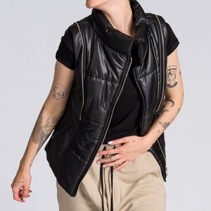 May include: A black puffer vest with gold zippers is worn over a black t-shirt and tan drawstring pants. The vest has a high collar. The person is smiling and has tattoos on their arms.