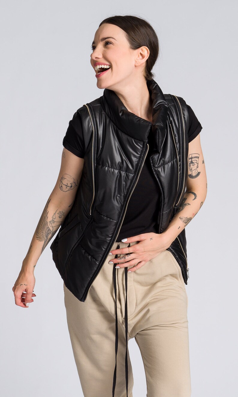 May include: A black puffer vest with gold zippers is worn over a black t-shirt and tan drawstring pants. The vest has a high collar. The person is smiling and has tattoos on their arms.