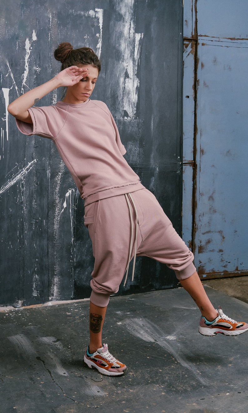 Op de afbeelding: Een vrouw draagt een roze sweatshirt en een bijpassende broek met een trekkoord. Ze draagt sneakers in wit, oranje en blauw.