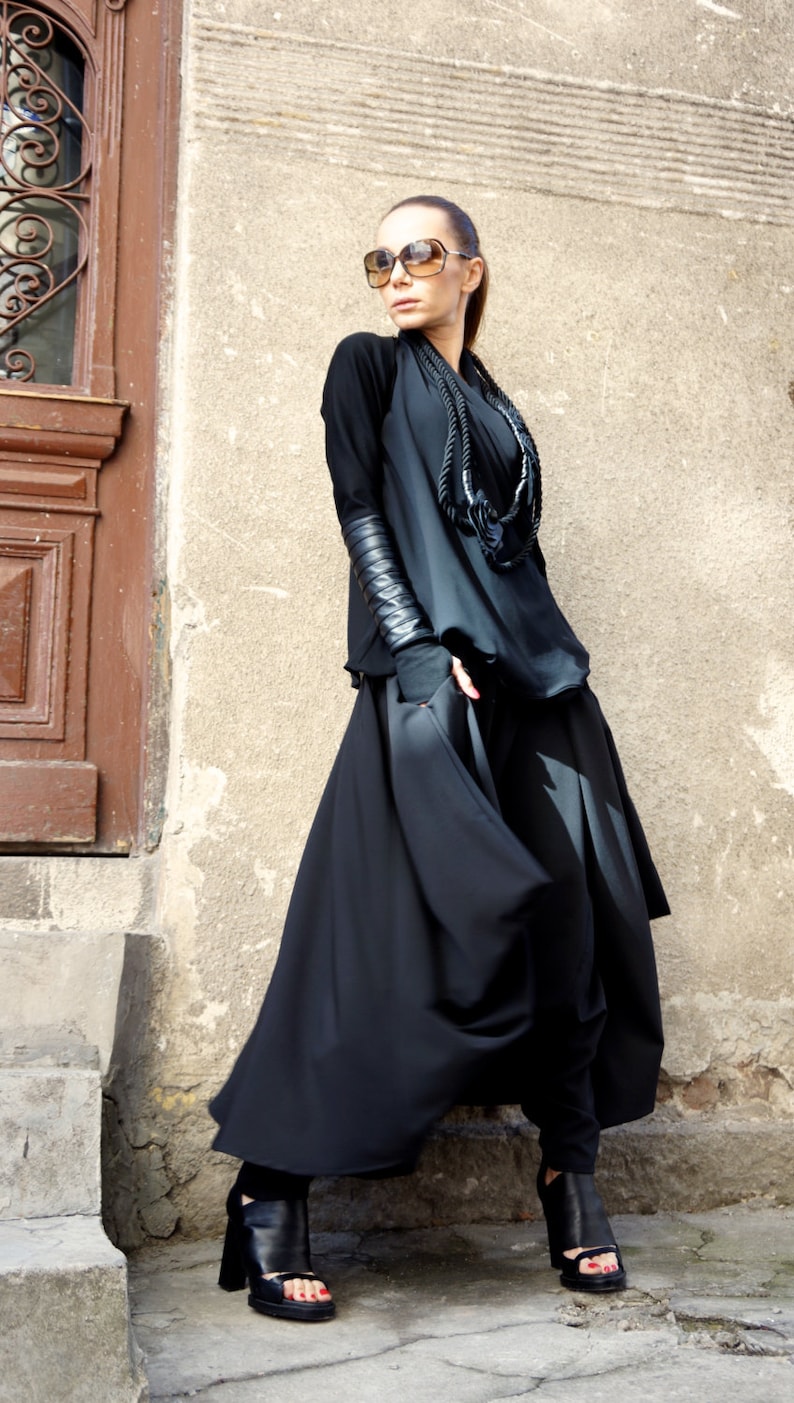 May include: A woman wearing a black long-sleeved top with leather accents on the sleeves, a black necklace, and a black skirt. She is wearing sunglasses and black heeled sandals.