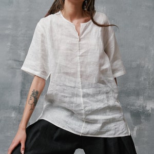 May include: A white linen shirt with a v-neck and short sleeves. The shirt is loose fitting and has a relaxed silhouette. The shirt is paired with black wide-leg pants.