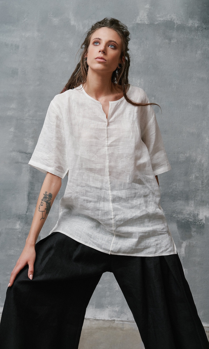 May include: A white linen shirt with a v-neck and short sleeves. The shirt is loose fitting and has a relaxed silhouette. The shirt is paired with black wide-leg pants.