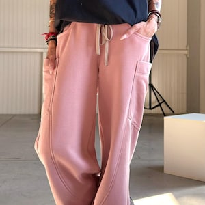 May include: A person is wearing a black t-shirt with white text and pink sweatpants with pockets. The sweatpants have a drawstring waist. The person is also wearing gray sneakers.