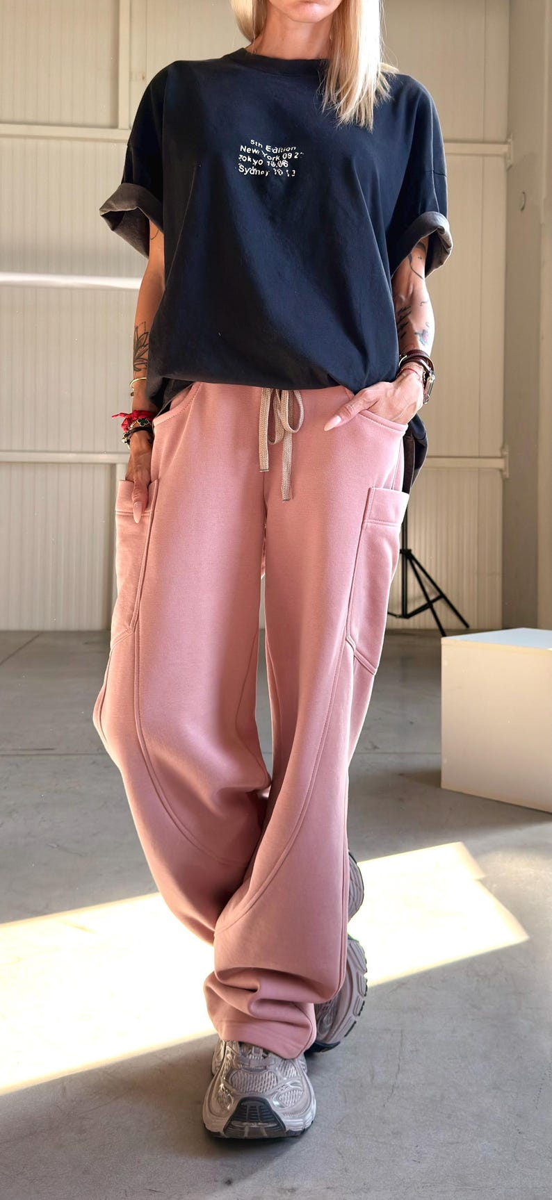 May include: A person is wearing a black t-shirt with white text and pink sweatpants with pockets. The sweatpants have a drawstring waist. The person is also wearing gray sneakers.