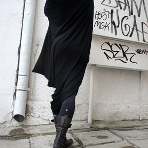 Op de afbeelding: Een vrouw draagt een zwarte, lange, gedrapeerde tuniek met leggings en bruine laarzen. De tuniek is losjes gesneden en heeft een vloeiende, asymmetrische zoom. De vrouw staat voor een witte muur met graffiti.