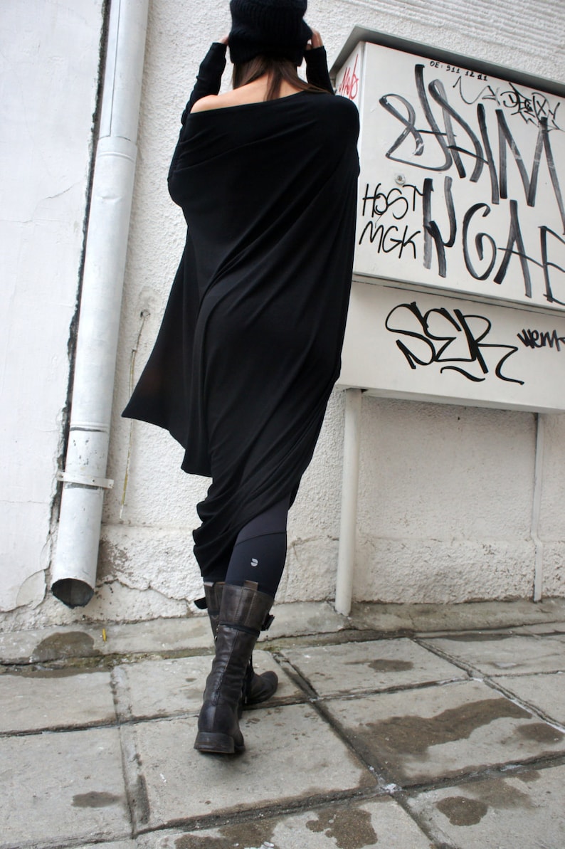 Op de afbeelding: Een vrouw draagt een zwarte, lange, gedrapeerde tuniek met leggings en bruine laarzen. De tuniek is losjes gesneden en heeft een vloeiende, asymmetrische zoom. De vrouw staat voor een witte muur met graffiti.