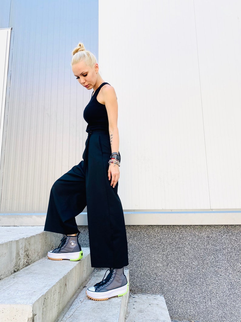 May include: A person wearing black wide-leg pants and a black tank top with a white belt. They are wearing black and grey high-top sneakers with a neon green accent.