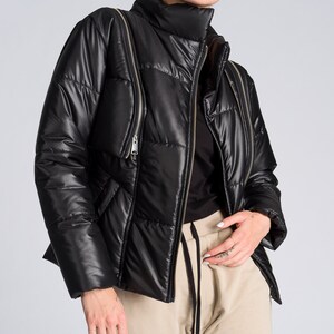 May include: Black puffer jacket with a high collar and multiple zipper details. The jacket has a quilted design and a shiny finish. The model is wearing a black top and tan sweatpants.