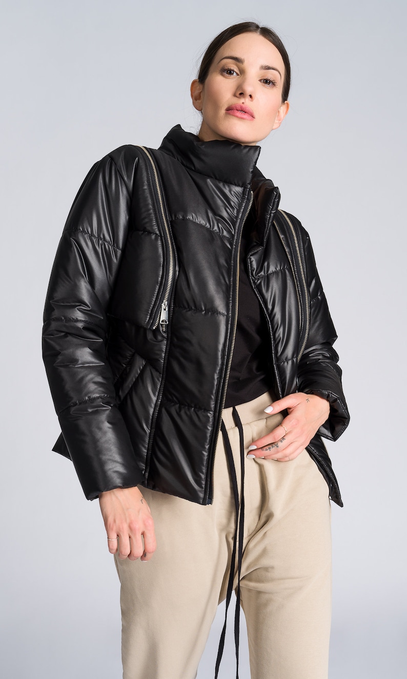 May include: Black puffer jacket with a high collar and multiple zipper details. The jacket has a quilted design and a shiny finish. The model is wearing a black top and tan sweatpants.