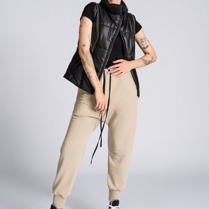 May include: A person wearing a black quilted vest with gold zippers over a black top, paired with beige sweatpants and high-top sneakers with black, white, and brown accents. The outfit is set against a plain white background.