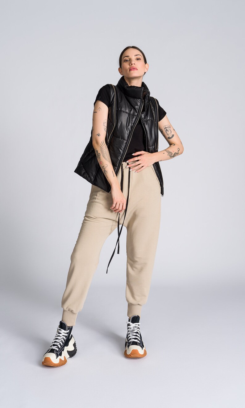 May include: A person wearing a black quilted vest with gold zippers over a black top, paired with beige sweatpants and high-top sneakers with black, white, and brown accents. The outfit is set against a plain white background.