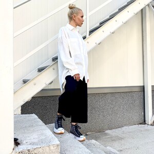 May include: A person wearing a white button-down shirt, black culottes, and black high-top sneakers with white soles. The person is standing on concrete steps next to a white metal staircase.