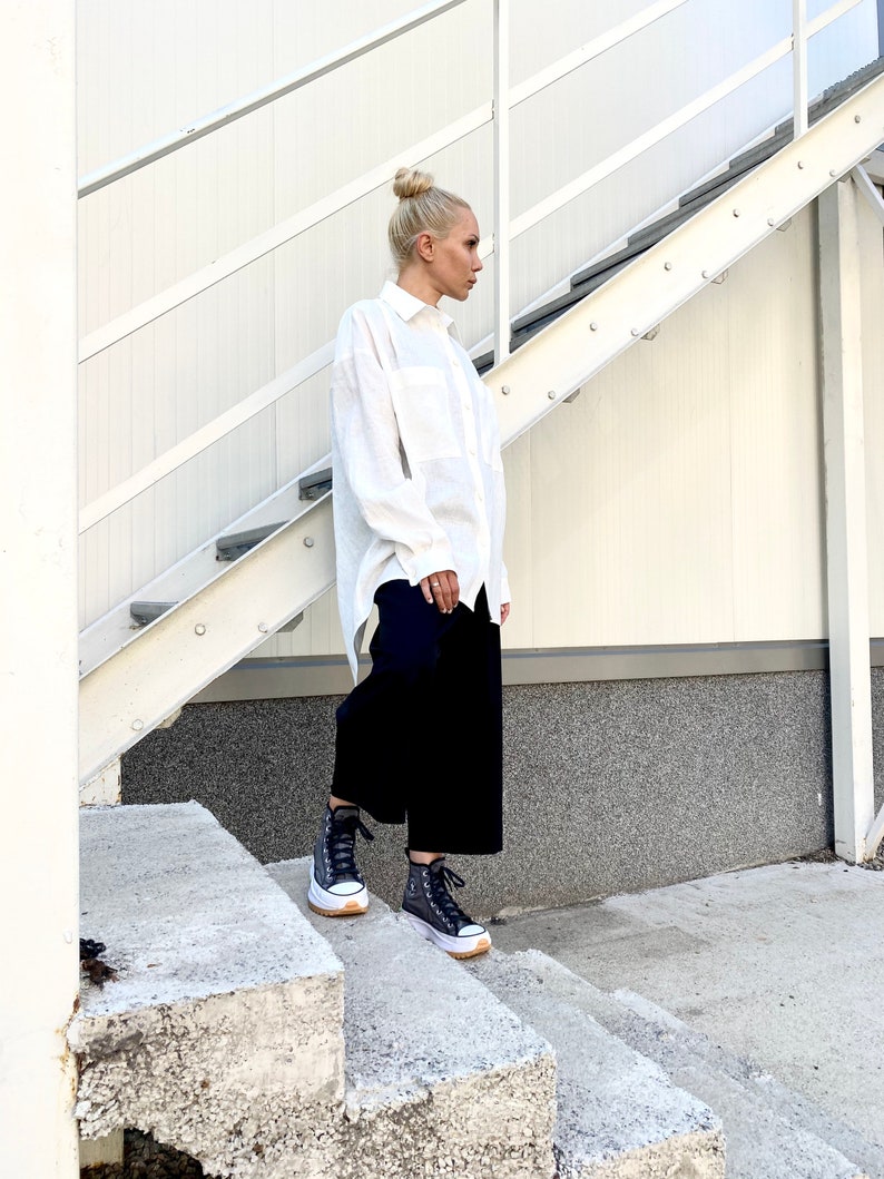 May include: A person wearing a white button-down shirt, black culottes, and black high-top sneakers with white soles. The person is standing on concrete steps next to a white metal staircase.