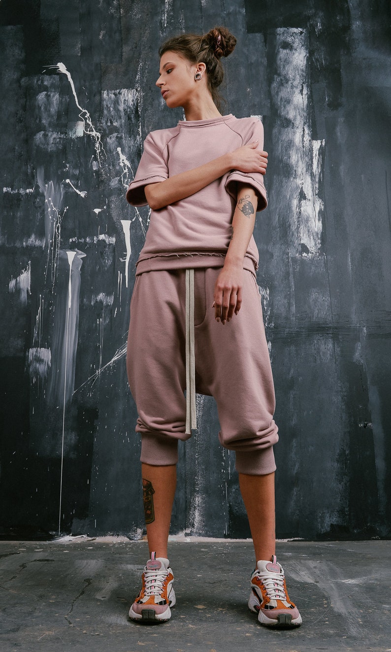 Op de afbeelding: Een vrouw draagt een stoffig roze sweatshirt en een bijpassende joggingbroek met een trekkoord. Ze draagt witte sneakers met oranje en bruine accenten.