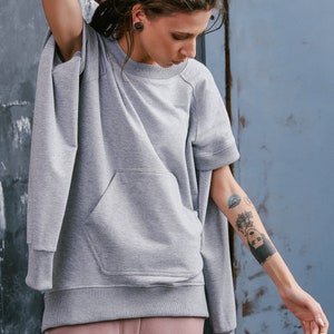 May include: A gray sweatshirt with a large pocket on the front. The sweatshirt has a relaxed fit and short sleeves. The model is wearing the sweatshirt with pink sweatpants.