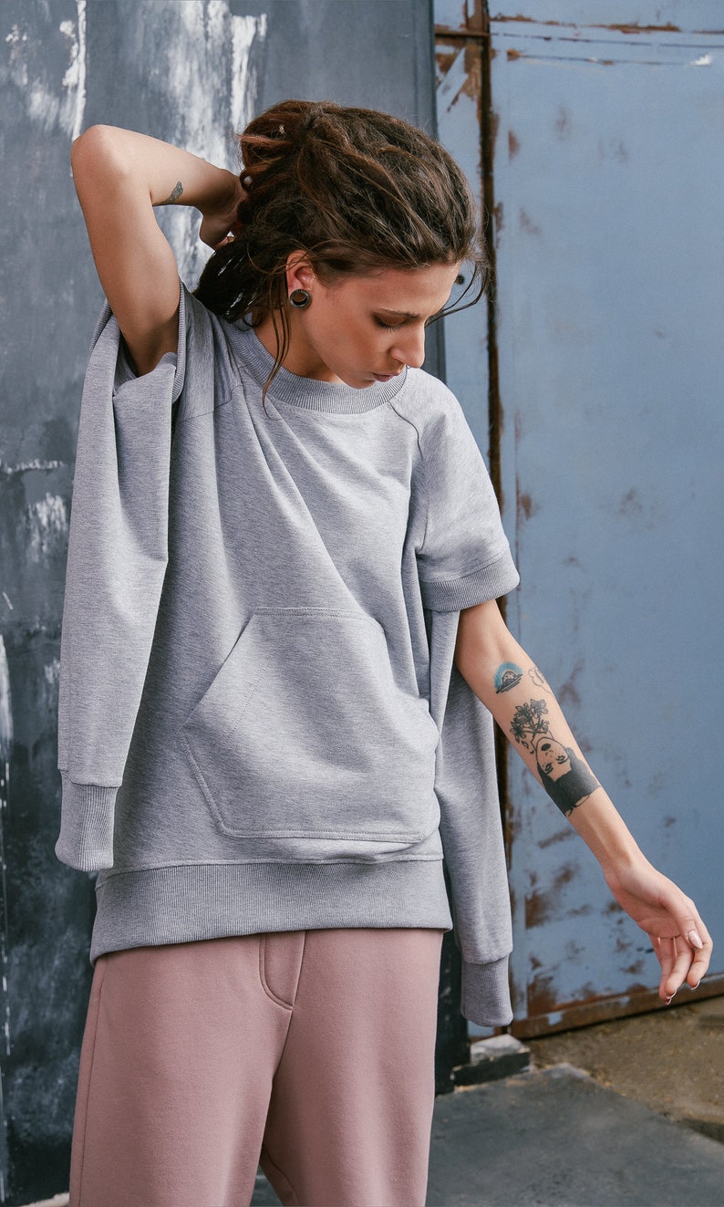 May include: A gray sweatshirt with a large pocket on the front. The sweatshirt has a relaxed fit and short sleeves. The model is wearing the sweatshirt with pink sweatpants.