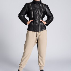 May include: Black puffer jacket with multiple silver zippers and a high collar. The jacket is paired with beige sweatpants and high-top sneakers with black, white, and brown accents. The outfit is set against a light gray background.