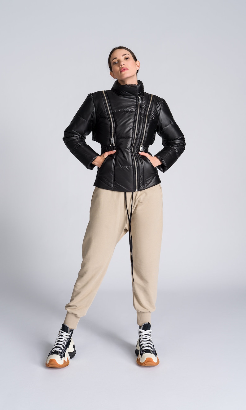 May include: Black puffer jacket with multiple silver zippers and a high collar. The jacket is paired with beige sweatpants and high-top sneakers with black, white, and brown accents. The outfit is set against a light gray background.