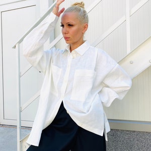 May include: A white linen shirt with two front pockets and long sleeves. The shirt is unbuttoned, revealing black pants. The model is standing in front of a white wall and stairs.