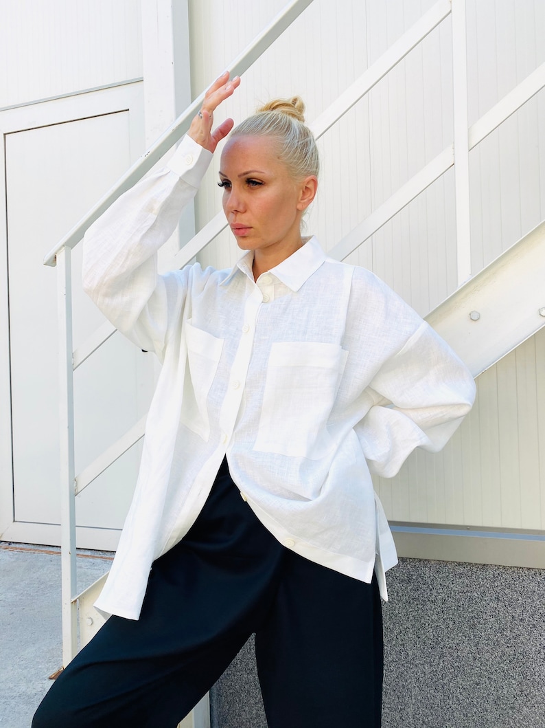 May include: A white linen shirt with two front pockets and long sleeves. The shirt is unbuttoned, revealing black pants. The model is standing in front of a white wall and stairs.