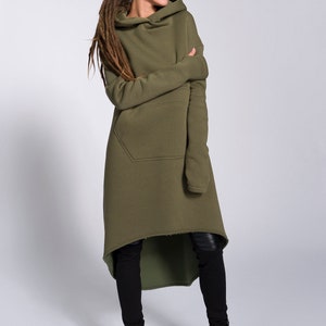 May include: A woman wearing a long, olive green hooded sweatshirt with a high neckline and a single pocket. The sweatshirt has a high-low hemline and is styled with black leggings and black sneakers.