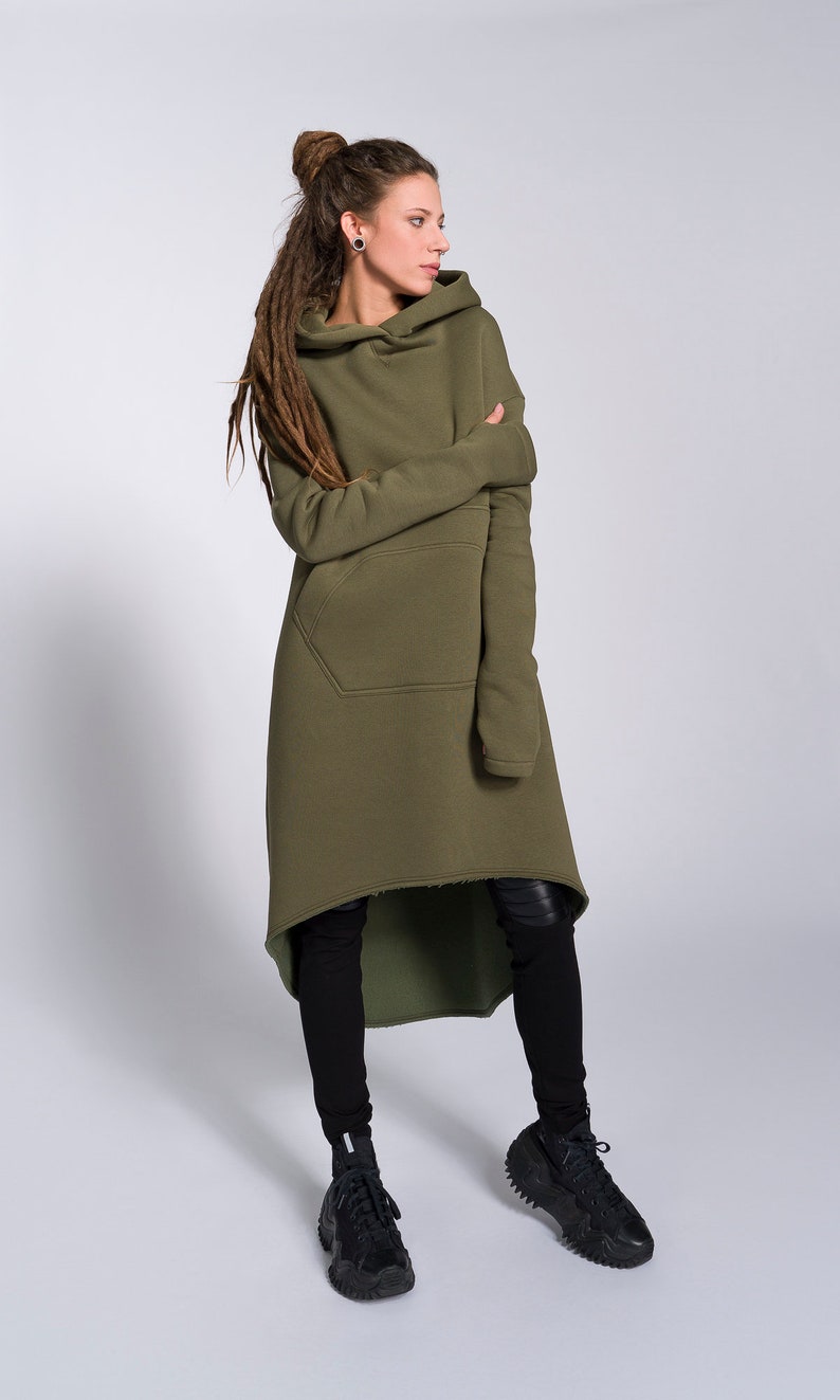 May include: A woman wearing a long, olive green hooded sweatshirt with a high neckline and a single pocket. The sweatshirt has a high-low hemline and is styled with black leggings and black sneakers.