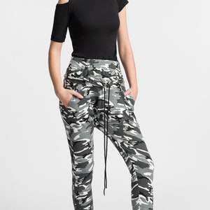 May include: A woman wearing a black short-sleeved shirt with a cut-out shoulder detail and camouflage print pants. She is wearing black high-top sneakers with white soles.