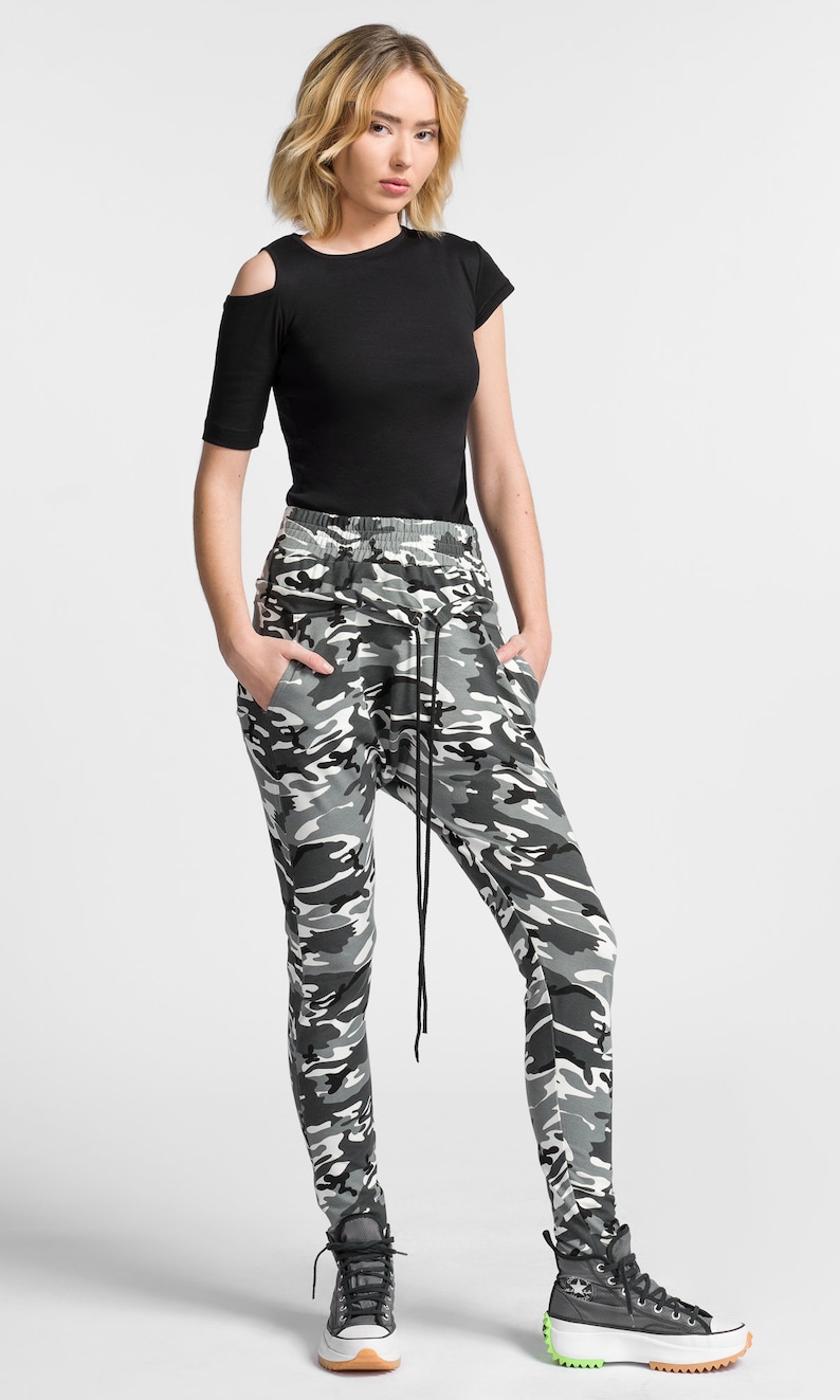 May include: A woman wearing a black short-sleeved shirt with a cut-out shoulder detail and camouflage print pants. She is wearing black high-top sneakers with white soles.