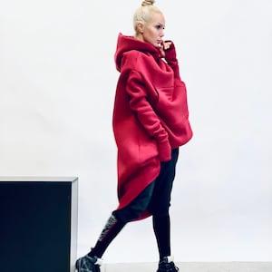 以下が含まれることがあります： 大きなフードと親指を通す穴のある袖口を備えた、オーバーサイズの赤い長袖パーカー。この衣服は、前後で長さの異なる裾とフロントポケットが特徴です。黒いパンツ、レギンス、白いアクセントの入った黒いスニーカーと組み合わされています。