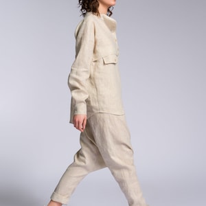 May include: A person wearing a light beige linen pantsuit with a collared shirt and long sleeves. The pantsuit has a relaxed fit and the pants are slightly flared at the bottom. The person is wearing white high-top sneakers with a light brown sole.