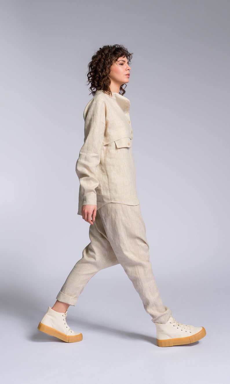 May include: A person wearing a light beige linen pantsuit with a collared shirt and long sleeves. The pantsuit has a relaxed fit and the pants are slightly flared at the bottom. The person is wearing white high-top sneakers with a light brown sole.