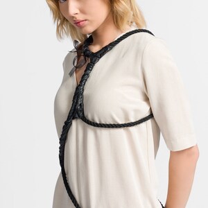 May include: A woman wearing a white linen dress with a black rope harness. The rope is tied in a knot at the neck and wraps around the chest and waist.