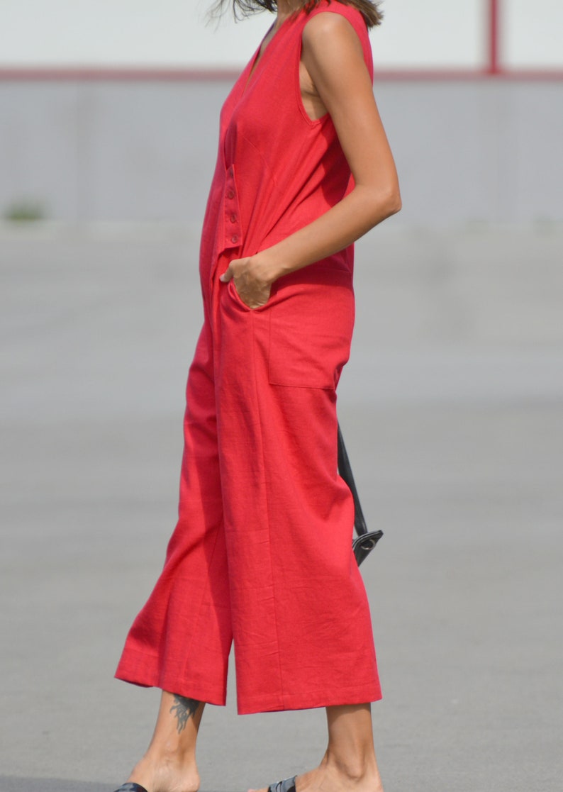 May include: A woman wearing a sleeveless red jumpsuit with a V-neckline. The jumpsuit has a button closure and pockets.