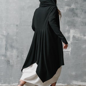 May include: A black hooded long-sleeved top with a flowing, asymmetrical hemline. The top is worn over a white skirt and paired with black and white trainers with red soles.