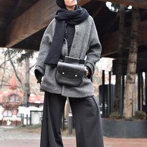 May include: A woman wearing a grey wool coat with a black knit scarf and a black leather bag. She is wearing black wide-leg trousers and black shoes.