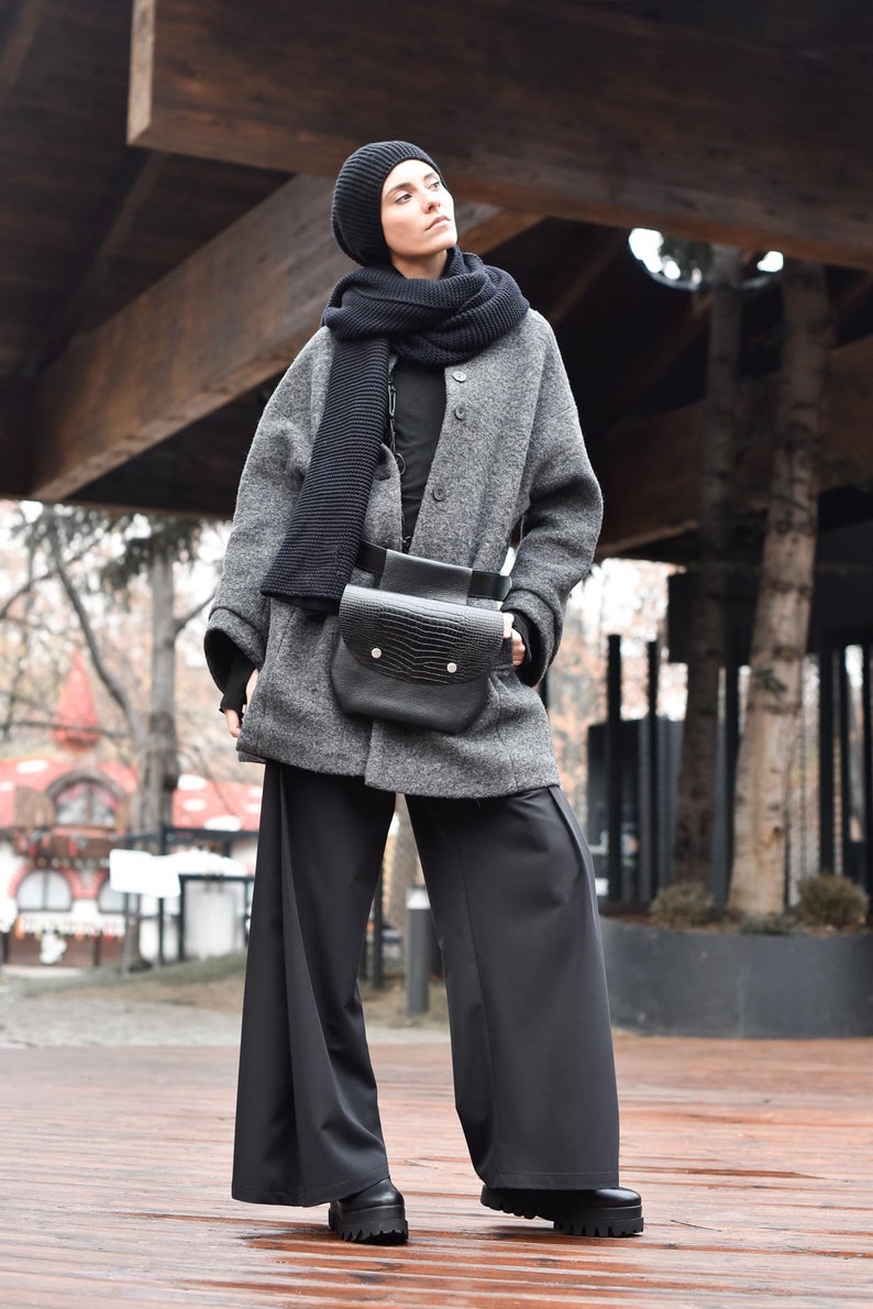 May include: A woman wearing a grey wool coat with a black knit scarf and a black leather bag. She is wearing black wide-leg trousers and black shoes.