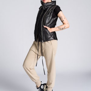 May include: Black quilted puffer vest with a high collar and a full-length zipper. The vest is worn over a black t-shirt and paired with tan joggers and black and white high-top sneakers with a chunky sole. The outfit is casual and modern.