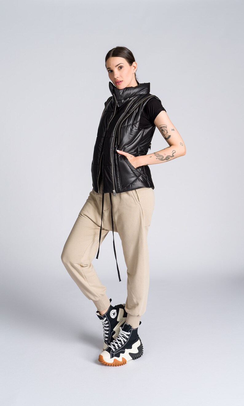 May include: Black quilted puffer vest with a high collar and a full-length zipper. The vest is worn over a black t-shirt and paired with tan joggers and black and white high-top sneakers with a chunky sole. The outfit is casual and modern.