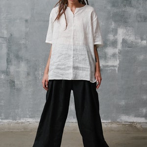 May include: A white linen shirt with a v-neck and short sleeves. Black linen pants with pleats. White sneakers with brown soles.