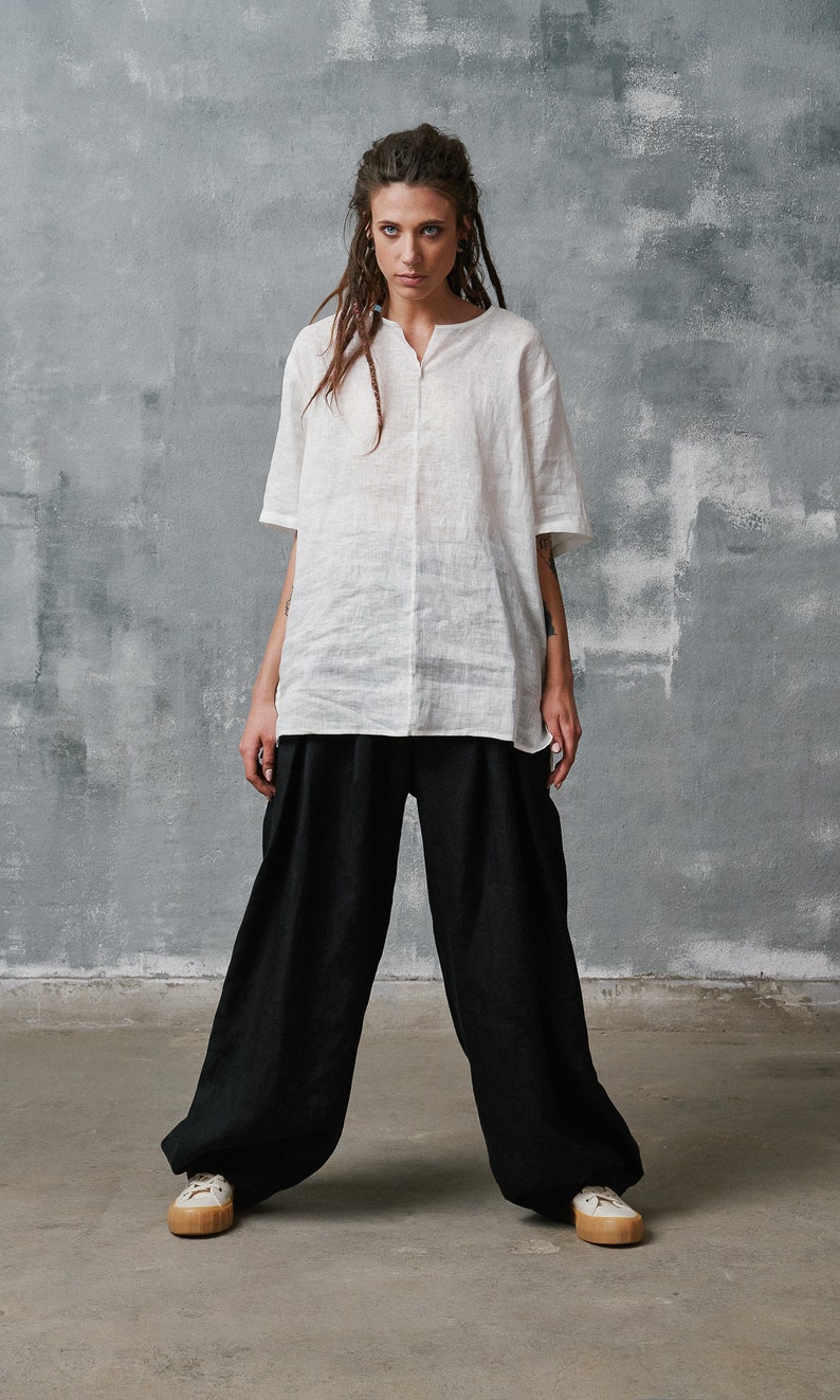 May include: A white linen shirt with a v-neck and short sleeves. Black linen pants with pleats. White sneakers with brown soles.