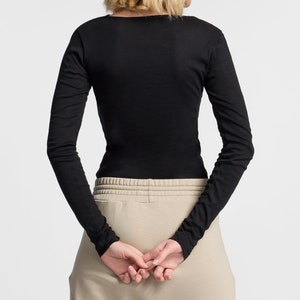 Puede incluir: Una camiseta negra de manga larga con cuello redondo y silueta ajustada. La camiseta se lleva con pantalones beige.