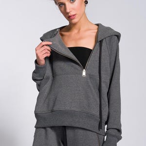 May include: A grey hooded sweatshirt with a half-zip and matching grey sweatpants. The sweatshirt features a front pocket and black drawstrings. The model is wearing a black tube top under the sweatshirt.