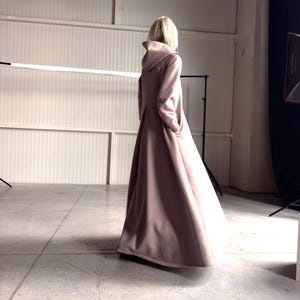 May include: A long, flowing, dusty rose-colored coat with a hood. The coat is floor-length and has a simple, elegant design. The model is standing in a studio setting, with a neutral background.